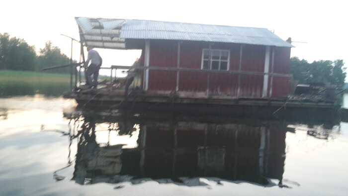 Röd husbåt med plåttak, delvis nedbruten, bogserad på en lugn sjö, person står på däck vid solnedgång.