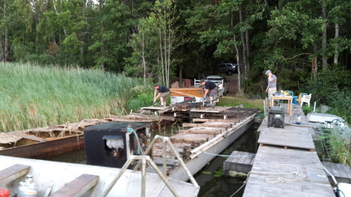 Personer demonterar en gammal husbåt som ligger vid en brygga omgivet av träd och gräs.
