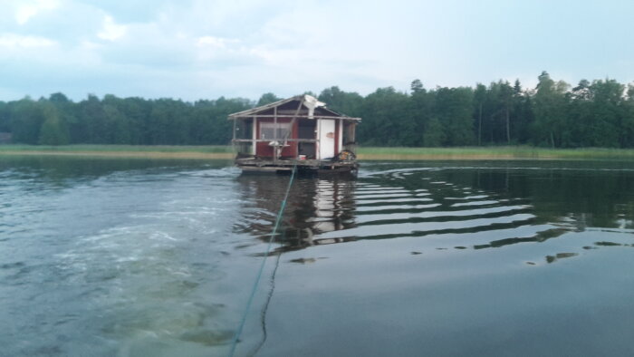 Husbåt bogseras över vattnet med en lina, omgivna av skog, för att nå sin sista viloplats.