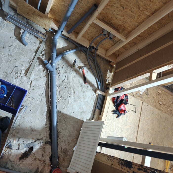 Byggplats med installerade grå rör i en vägg och på golv; synligt verktyg och arbetsmateriel.