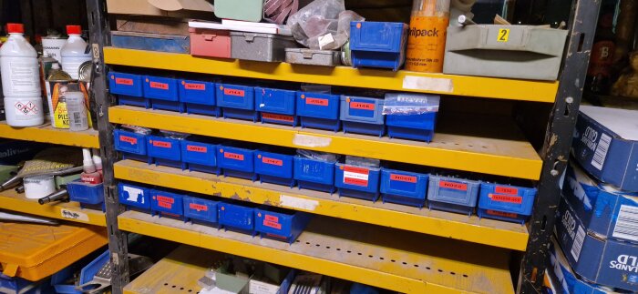 Gula hyllor med flera blå plastlådor märkta med olika koder, omgivna av verktyg och flaskor i en verkstadsmiljö.