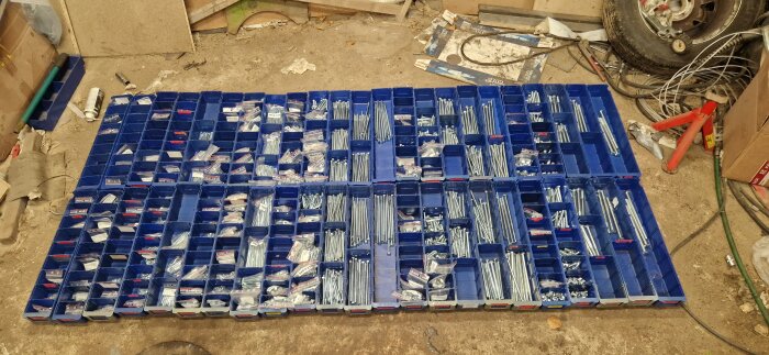 Blå lådor fyllda med olika skruvar och muttrar sorterade i ett garageutrymme. Verktyg och bildelar syns i bakgrunden.