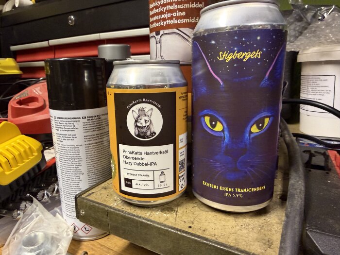 Två ölbokar, en med etiketten "PrinsKatts Hantverksöl" och en med en kattbild. Bakgrund med verktyg och sprayflaskor.