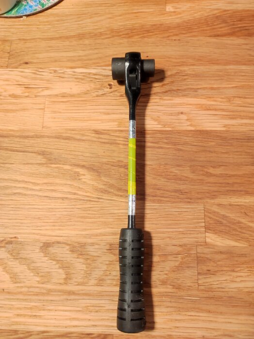 Svart och gult handverktyg med hammarliknande huvud, hittat i en verktygslåda, liggande på ett trägolv. Märke: Super, Made in Japan.