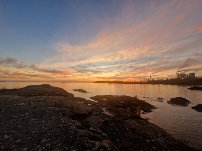 Solnedgång över sjön Vänern med klippor i förgrunden och silhuetten av ett industriområde i bakgrunden.