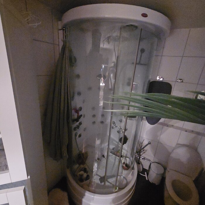 Rund ångduschkabin med glasdörrar och flera duschmunstycken, placerad i ett badrum med vita kakelväggar och toalett bredvid.