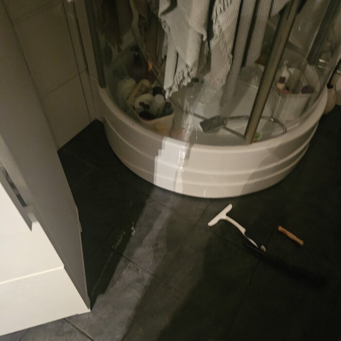 Duschkabin med vatten på golvet bredvid, duschskrapa och handduk synliga.