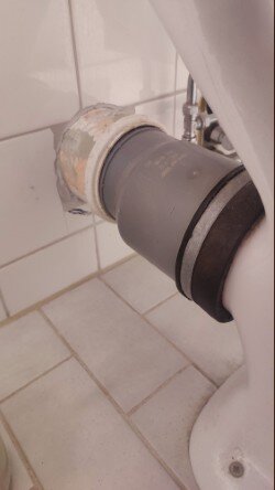 Toalettstol med P-lås ansluten till ett avloppsrör i ett litet badrum. Rören är sammankopplade med en grå manschett.