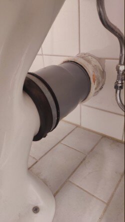 Toalettanslutning med grått avloppsrör och manschett i ett litet badrum, fotograferad från sidan, visar vattenrör och vita kakelplattor.