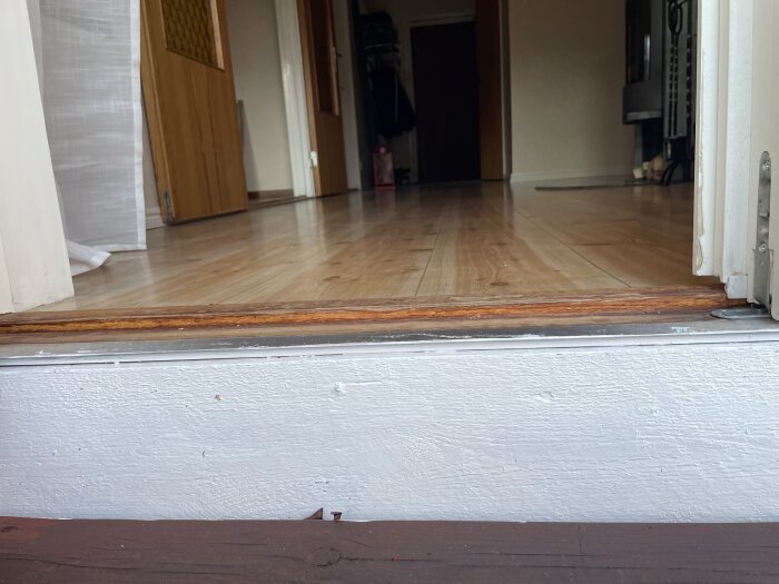 Altandörrens tröskel utan plåt, med vy in i huset över ett ljust trägolv och delvis öppna dörrar.