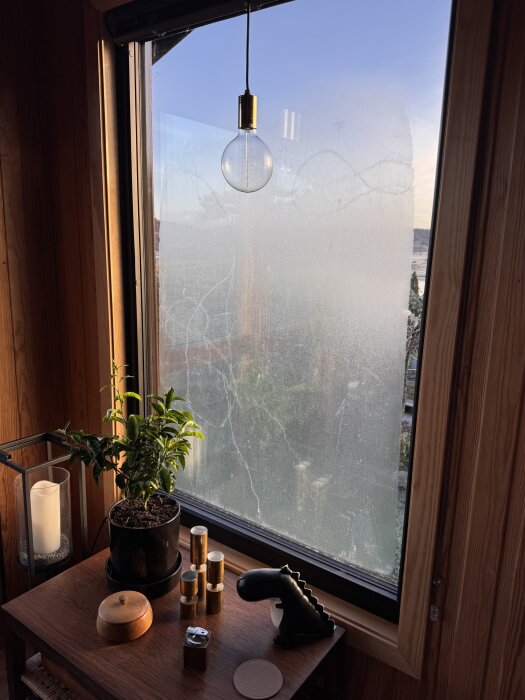 Fönster med imma och frost i hörn, inomhus perspektiv med bord, krukväxt och inredningsdetaljer.