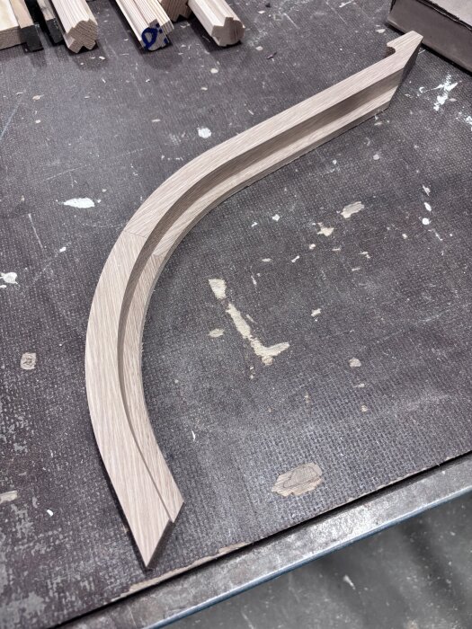 Böjd falsad kantlist i trä redo för limning på arbetsbänk, omgiven av andra bearbetade träbitar.