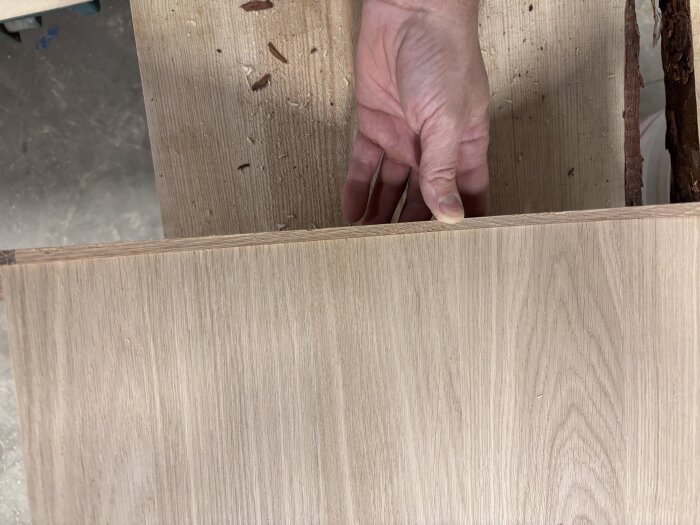 En hand håller en träbit med falsad kant, redo för limning. Bitar av sågspån syns på arbetsbänken.