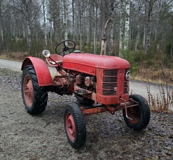 En röd Volvo T21 traktor står på en grusväg, omgiven av höstliga träd i bakgrunden.