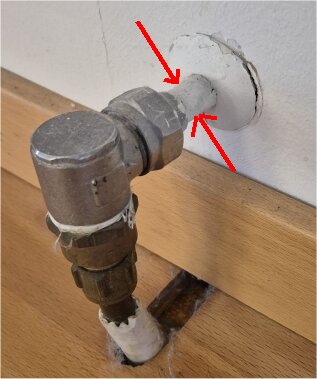 Koppling av metallrör genom vitmålad vägg med två röda pilar markerade, visar detaljer av en äldre ventilanordning inomhus.