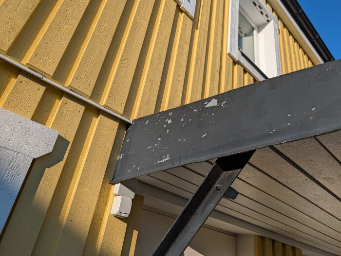 Grått entrétak med flagnande färg mot en gul träfasad; diskuteras underhållsmetoder för galvaniserat material.