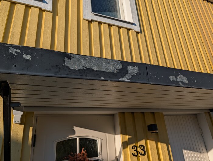 Galvat entrétak med flagande färg över en vit dörr på ett gult hus.