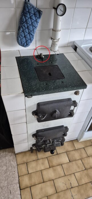 Gammal vedspis med termometer på rör. En del av vedspisen är inringad. Hängare med grytlapp syns bredvid.