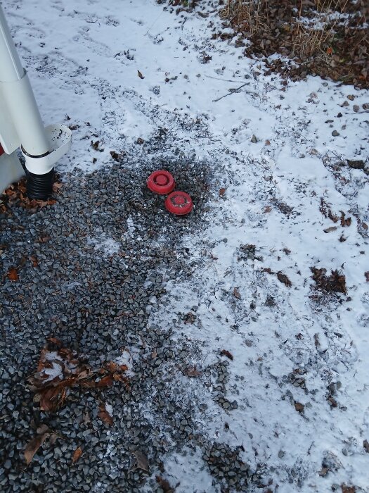Två röda rör med lock i snö på en grusplan, nära ett stuprör, diskuterade som eventuella rensrör eller tilluft.