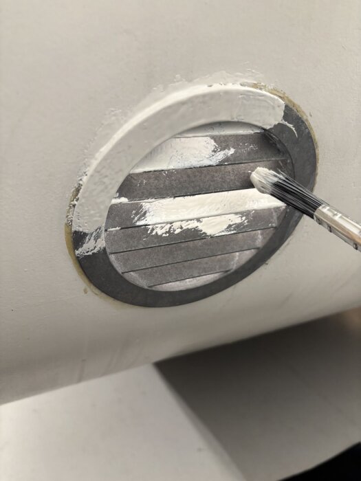 Runt ventilationsgaller på vit vägg målas med pensel och vit färg för att ta bort oxidation och ge en jämn finish.