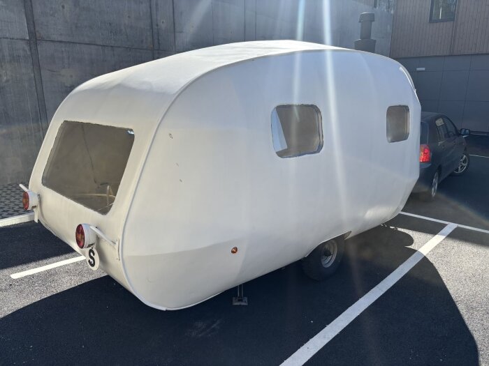 Vit husvagn parkerad på asfalt, sett bakifrån, med runda baklampor och sidofönster. En bil står bredvid, och husvagnen är del av renovering.