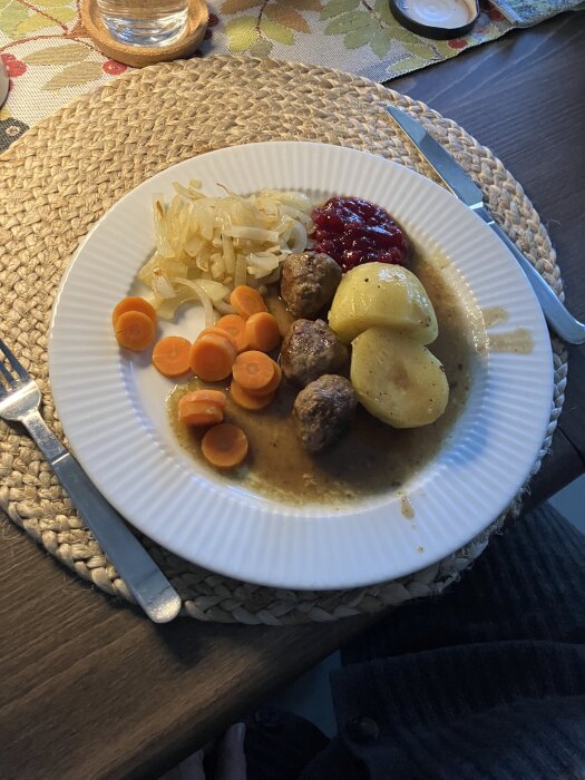 Tallrik med köttbullar, sås, potatis, lingonsylt, kokta morötter och stekt lök, serverat på en bordstablett av rotting.