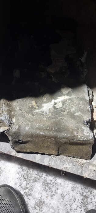 Hårt material under betonggolv i badrum, potentiellt asbest enligt inlägget, med sprickor och vitaktiga fläckar. En svart sko syns i vänstra hörnet.