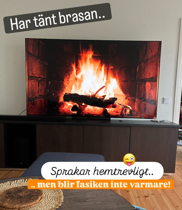 TV-skärm visar en sprakande brasa i en vardagsrumsmiljö, med texten "Har tänt brasan... Sprakar hemtrevligt.. men blir fasiken inte varmare!".