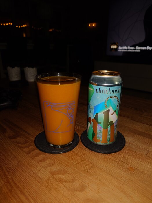 En ölburk märkt "elmeleven" med smak av mango och banoffee bredvid ett glas med orangefärgad dryck på ett bord.