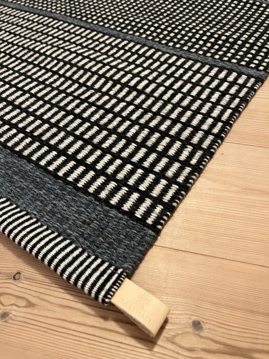 Svartvit mönstrad Kasthall Häggå ullmatta på trägolv, med mönster av horisontella och vertikala linjer.