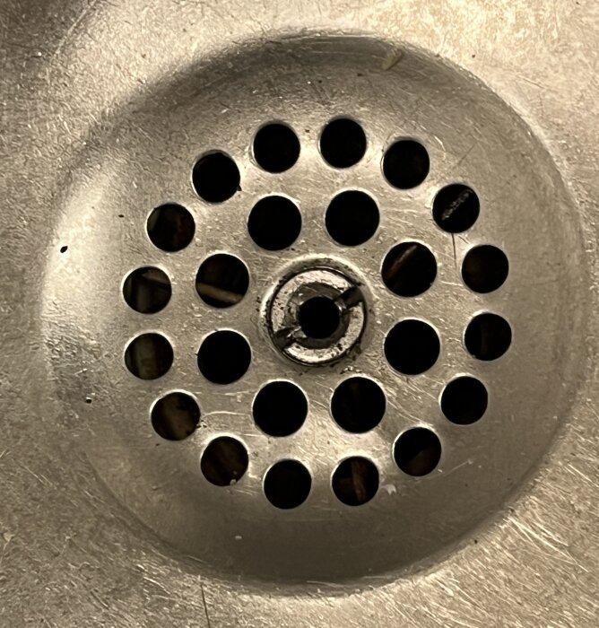 Diskbänksavlopp med bottenventil och skruv som verkar fastna, omgiven av hål i metall.