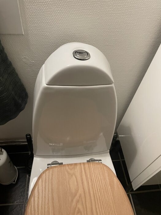 Toalett med träfärgat lock och dubbelsplushandtag på toppen, fotograferad ovanifrån.