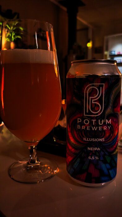 Ölglas med fyllig, bärnstensfärgad dryck bredvid en burk Potum Brewery Illusions NEIPA i dämpad inomhusbelysning.