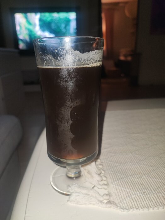 Ett glas med mörk hemmagjord black IPA på ett bord, bakgrund med suddig TV och möbler.