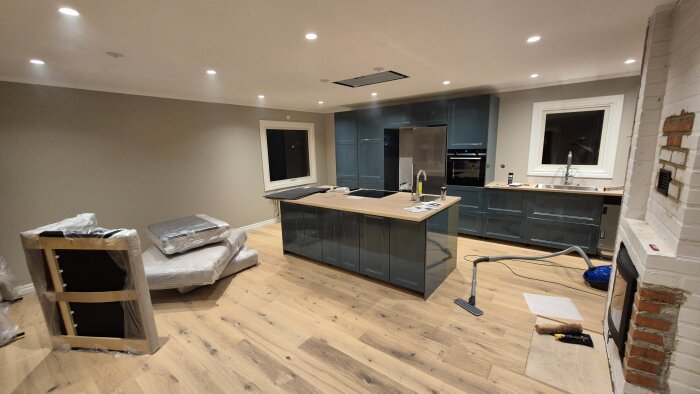 Nytt kök med köksö, ljusa trägolv och vitmålade väggar under renovering, möbler inslagna i plast, dammsugare på golvet.