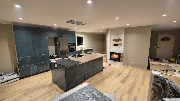 Modernt kök och vardagsrum under renovering med trägolv, blåa skåp, köksö och tänd spis.