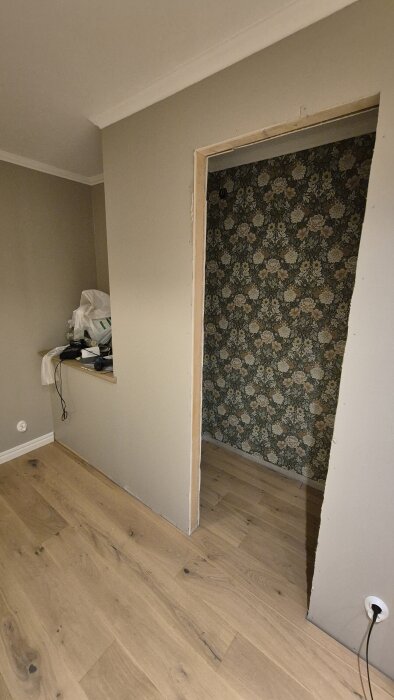 Öppet hål i väggen med blomtapet i ett rum under renovering, beige vägg och trägolv med saker på hylla.
