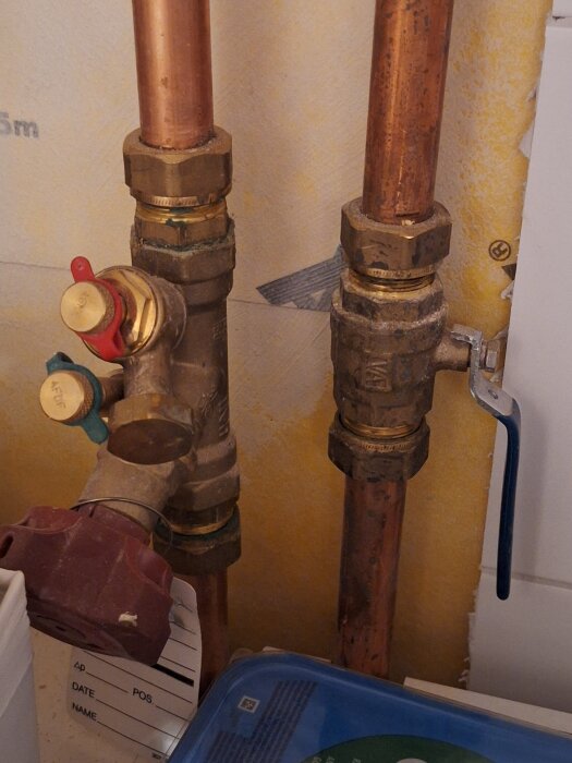 Kopparrör med ventiler, inklusive en röd kran, i ett system för radiatorer. Möjlig funktion för att reglera värmeflöde till övervåningen.