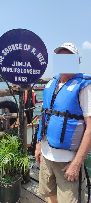 Person i flytväst vid skylten 'The Source of R. Nile, Jinja, World's Longest River' med vatten och båt i bakgrunden.