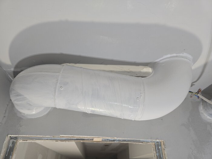 Vitmålad ventilationsrör i taket med ojämn yta, synlig koppling och synliga kablar bredvid rörändan.