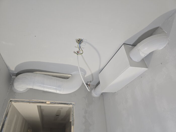 Vitmålad vägg och tak med synliga ventilationsrör och elledningar i ett renoveringsprojekt, innan ljuskällorna är installerade.