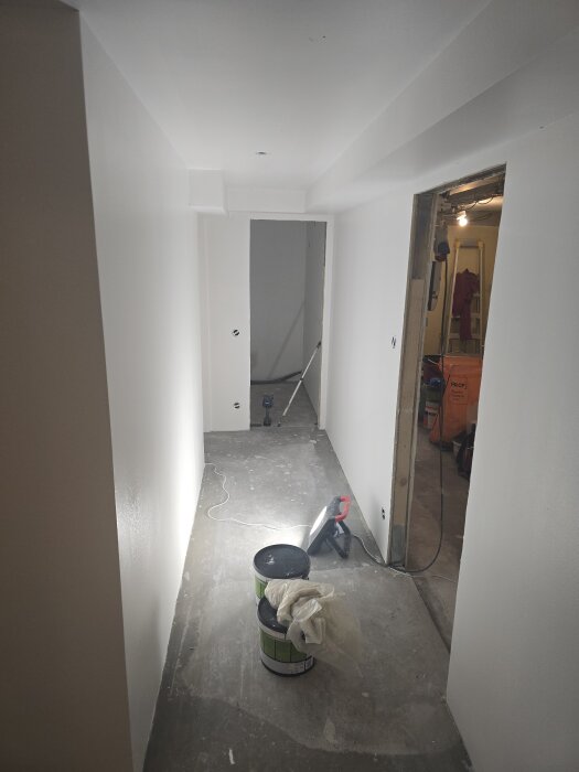 Nymålad vit korridor med oavslutat golv. Färgpytsar, verktyg och kablar syns längs väggarna. Öppen dörr till intilliggande rum.