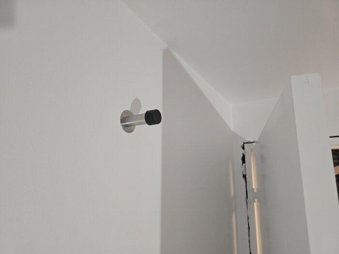 Dörrstop i metall monterat på nymålad vit vägg, i hörnet där två väggar möts. Dörren är delvis öppen.