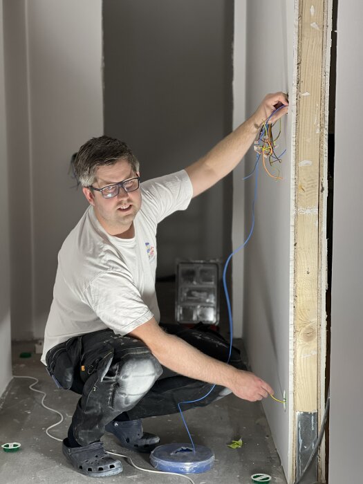 En person installerar elektriska ledningar i en nymålad korridor. Personen bär skyddsglasögon och arbetar med synliga kablar vid en väggöppning.