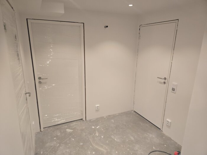 Nymålad vit korridor med två dörrar, plastade handtag och slätt grått golv; strömbrytare och elkontakt syns på väggen.