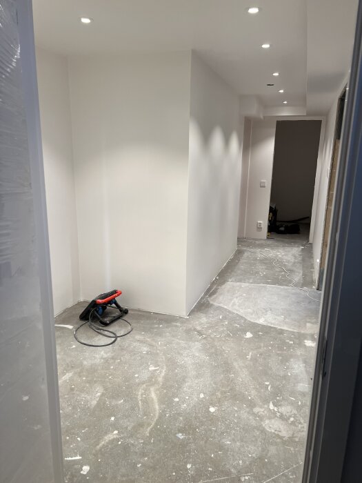 Nymålad korridor med vita väggar och infällda takspotlights, färdig för golvläggning. En stående lampa och några verktyg syns på golvet.