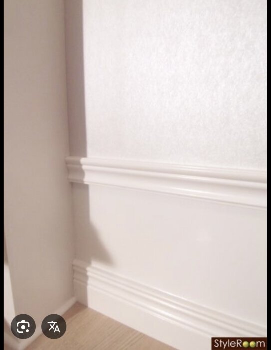 Vit vägg med golvsockel och bröstlist i allmogestil, där sockeln sitter ovanpå en bakre skiva. Listerna skapar en dekorativ panel på väggen.