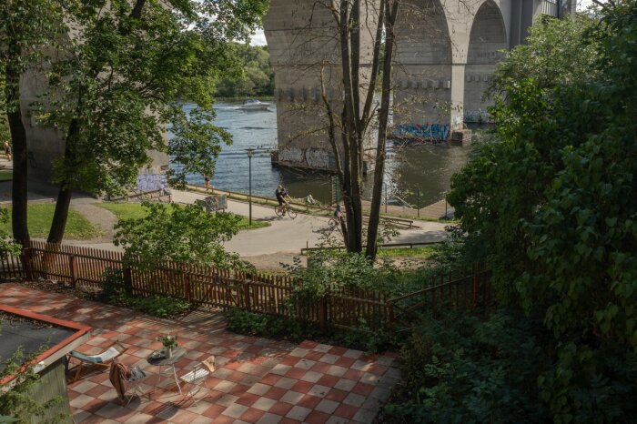 Utomhusplats med schackrutig plattläggning i ljusgrått och rödaktigt, belägen nära vatten och broar, omgiven av träd.