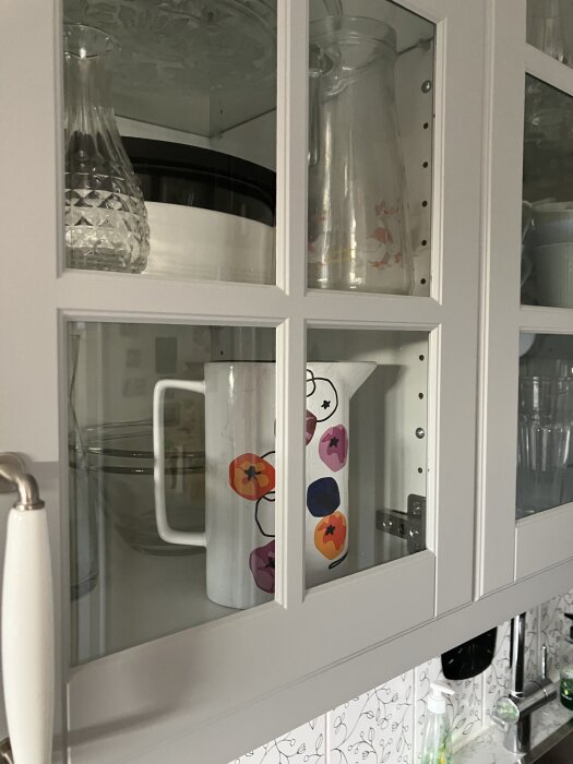 Skåp med glasdörrar visar karaffer och färgglad kanna. Synliga spikhuvuden på insidan av dörrkarmen.