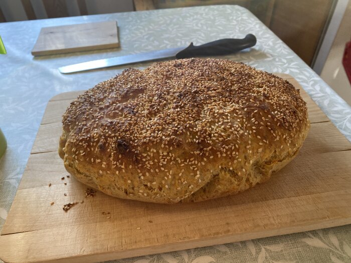 Sesamfröbröd på en skärbräda, bakat gyllenbrunt med ett brödkniv i bakgrunden på ett köksbord med duk.